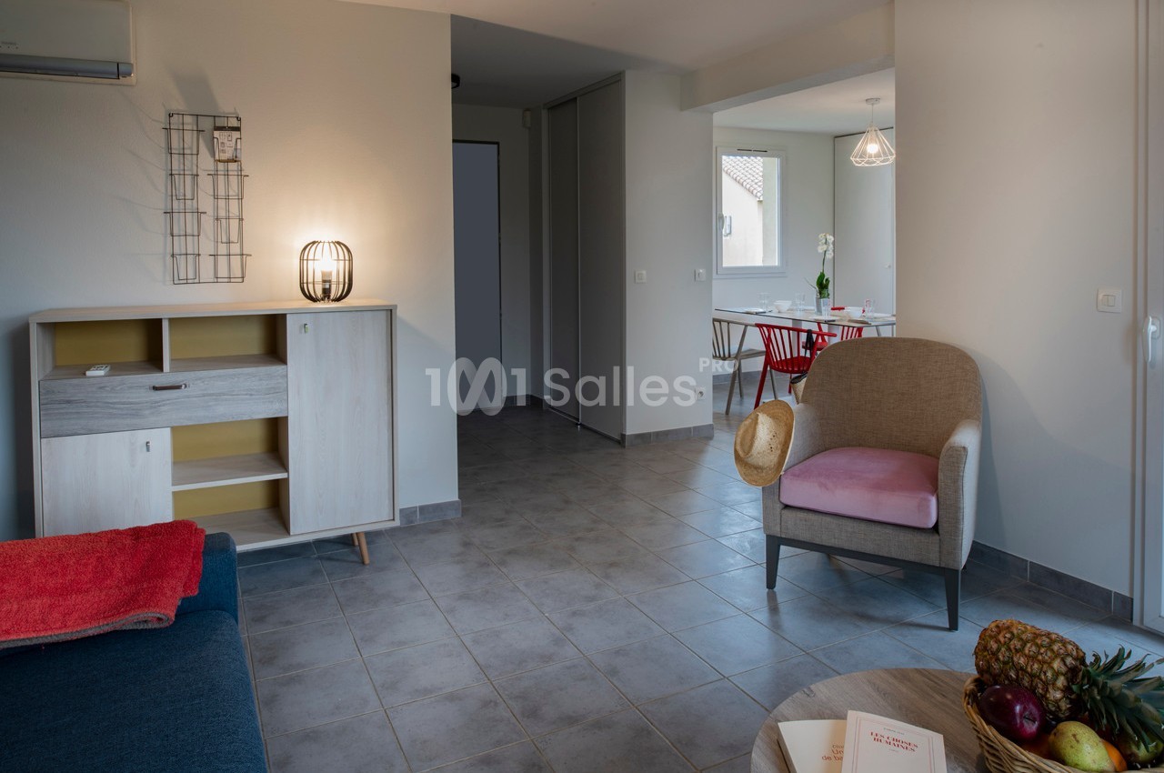 Salon lumineux avec mobilier moderne, sol en carrelage gris, chaise rose, buffet en bois et table à manger en arrière-plan.