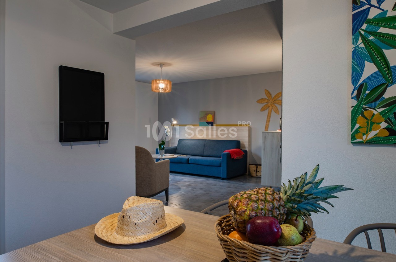 Vue intérieure d'une pièce lumineuse avec un salon en arrière-plan et une table décorée d'un panier de fruits au premier…