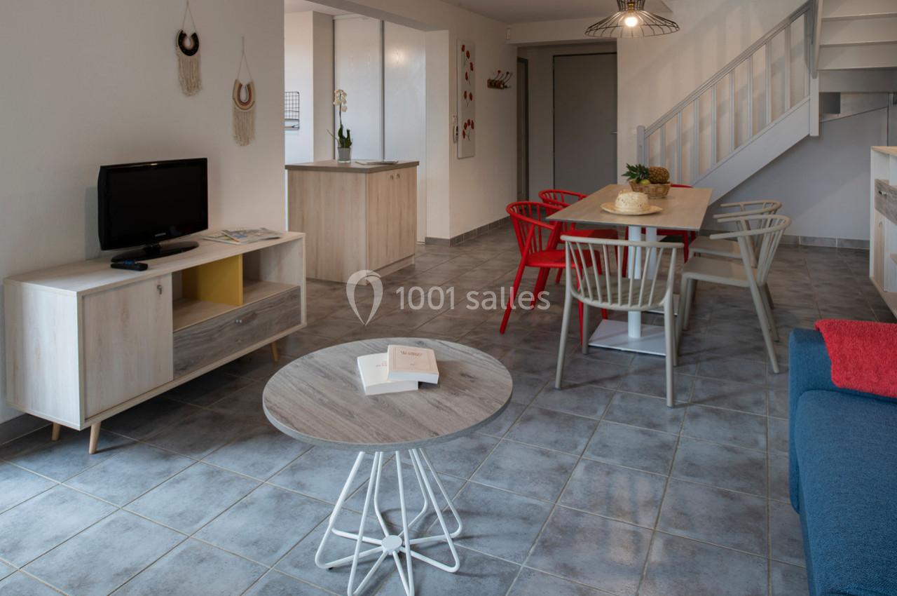 Salon moderne avec table à manger, chaises colorées, télévision, table basse et escalier dans un espace lumineux.
