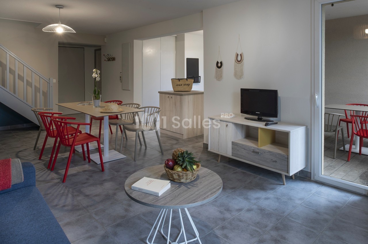 Salon moderne avec table à manger, chaises rouges, canapé, télévision et décoration murale, éclairé par une grande baie…