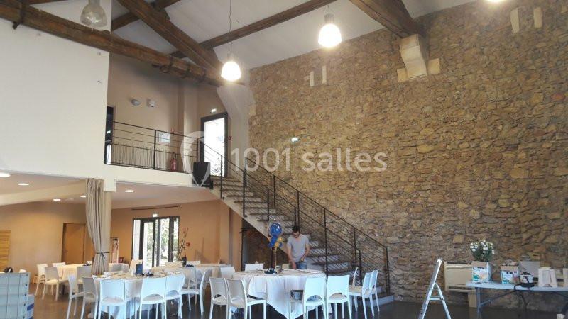 Salle de réception avec murs en pierre, tables dressées, escalier en métal et plafond avec poutres apparentes.