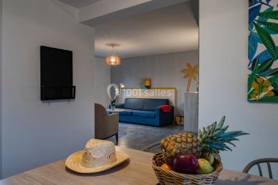 Salle à manger lumineuse avec table en bois, chaises colorées, cuisine ouverte et décoration sobre.