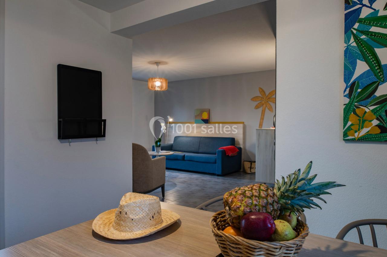 Vue d'un intérieur lumineux avec un salon en arrière-plan et une table avec un panier de fruits au premier plan.