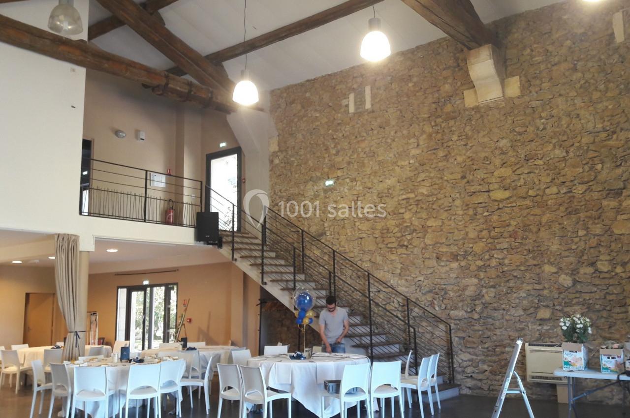 Salle avec murs en pierre, plafond à poutres apparentes, tables dressées et escalier métallique menant à une mezzanine.