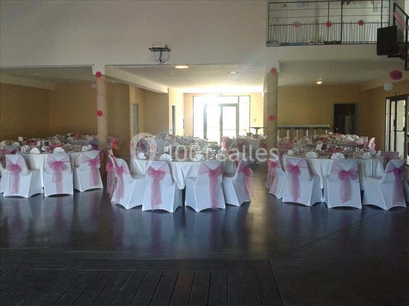 Salle de réception décorée avec des chaises blanches ornées de nœuds roses et des tables dressées pour un événement.
