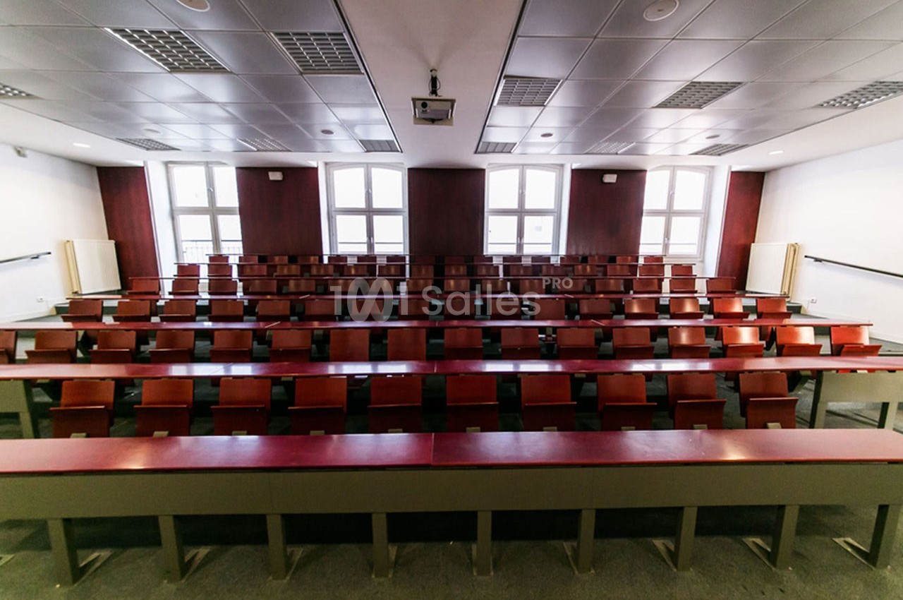 Salle de classe vide avec rangées de tables et chaises en bois, éclairée par des fenêtres et des lumières au plafond.