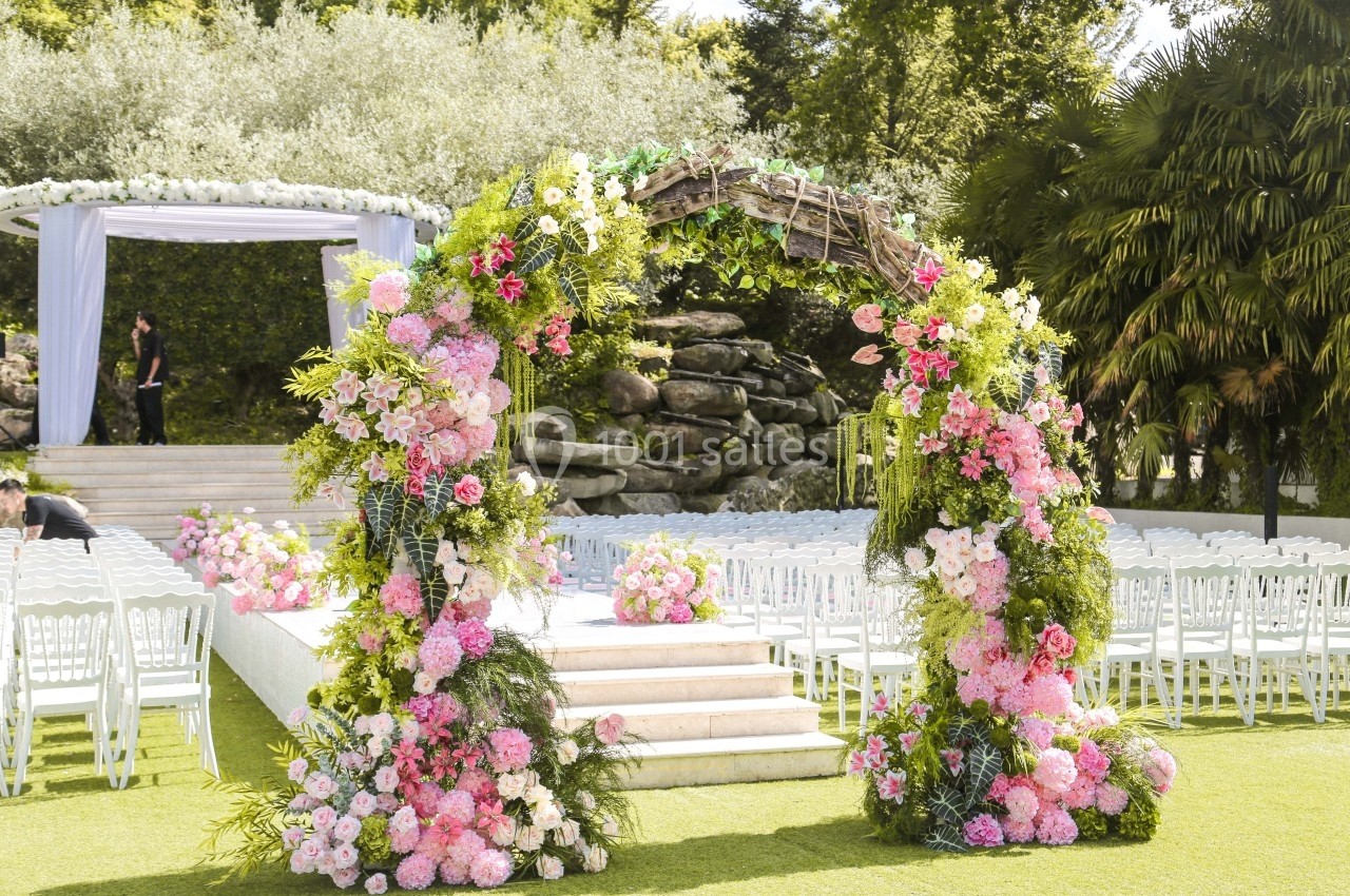 Arche florale ornée de roses et de feuillages devant des chaises blanches disposées pour une cérémonie en plein air.