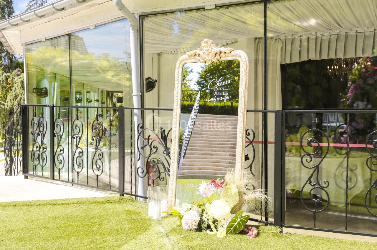 Grand miroir doré posé sur une pelouse, reflétant un escalier, entouré de fleurs et de bougies décoratives.