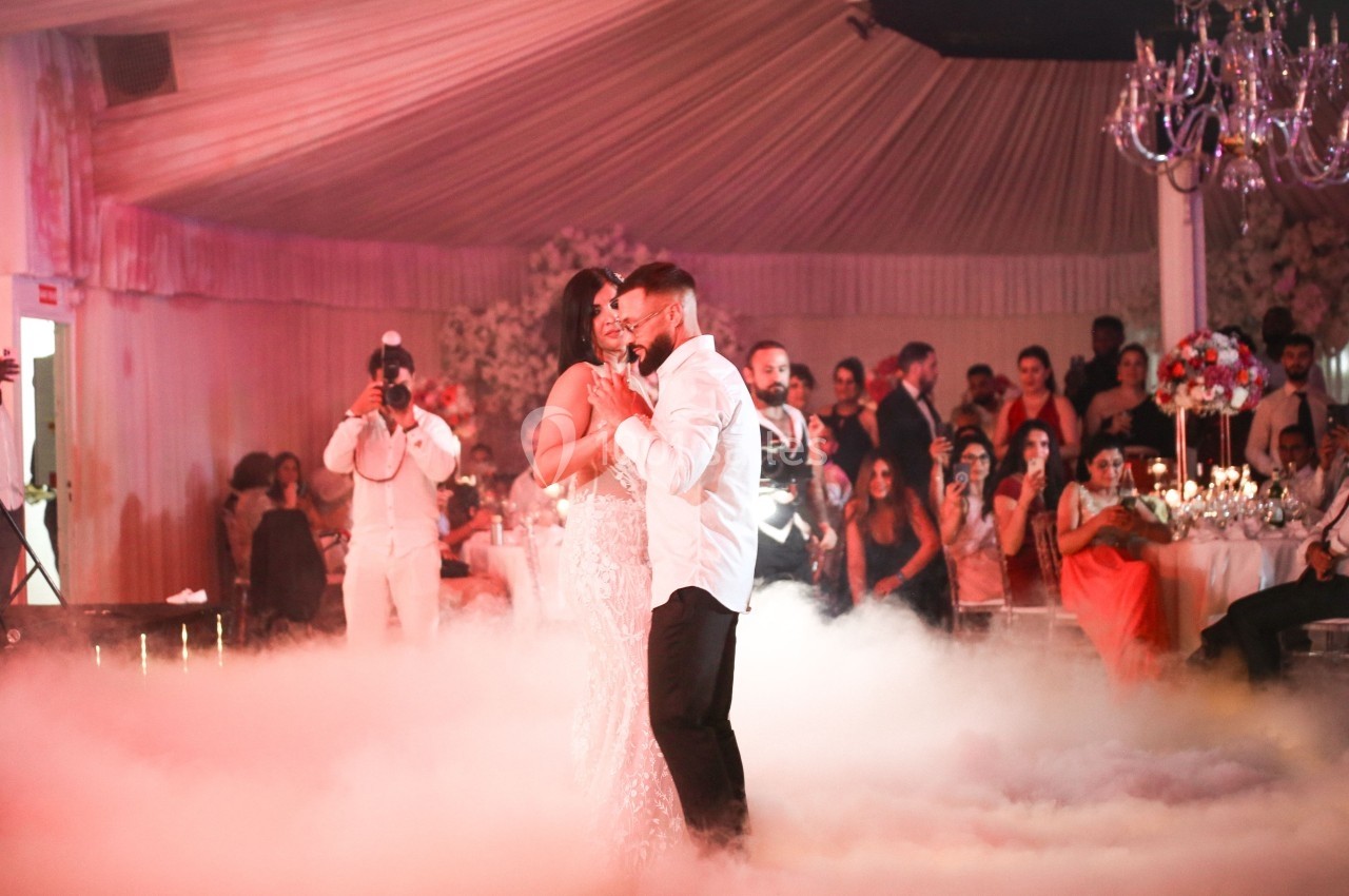 Un couple danse au centre d'une salle de réception décorée, entouré de fumée et d'invités observant la scène.