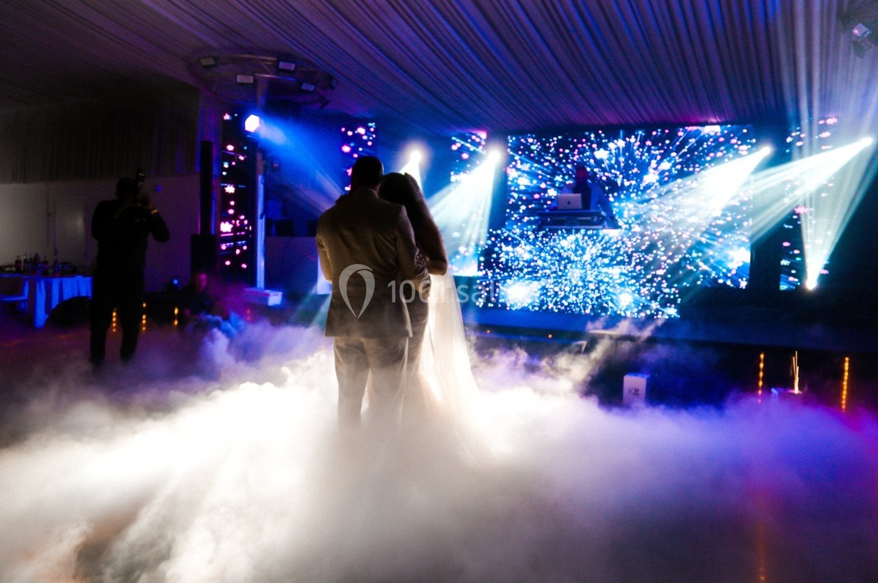 Un couple danse au milieu d'une piste enveloppée de fumée, éclairée par des projecteurs et un écran lumineux.