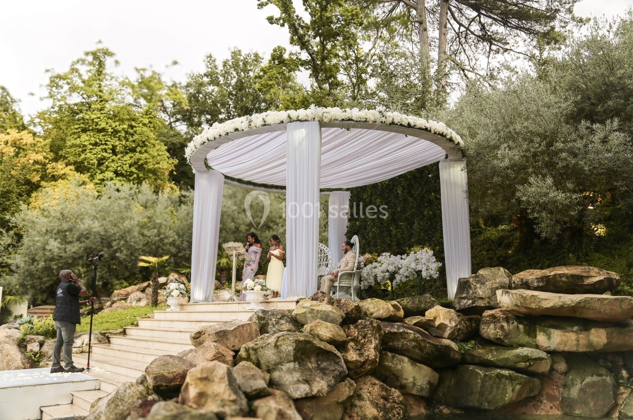 Un photographe capture une cérémonie sous une pergola décorée de fleurs blanches, entourée de verdure et de rochers.
