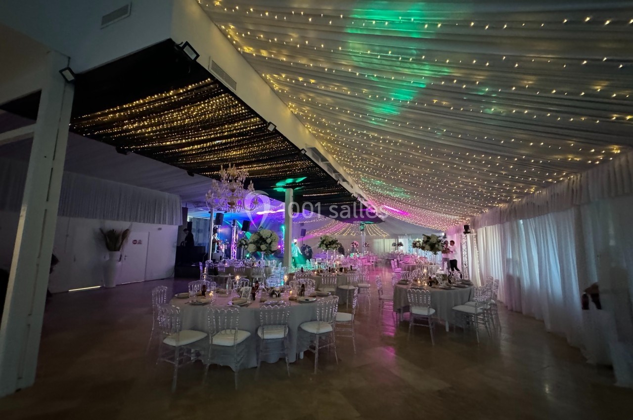 Salle de réception décorée avec des guirlandes lumineuses au plafond, tables dressées et éclairage coloré.