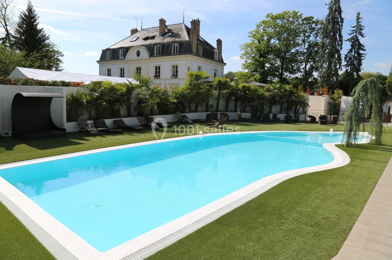 Piscine extérieure entourée de transats, pelouse synthétique et végétation, avec une grande maison en arrière-plan.