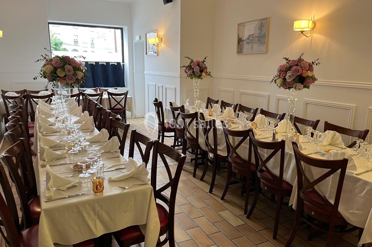 Salle de réception lumineuse avec deux longues tables dressées, décorées de fleurs et nappes blanches.
