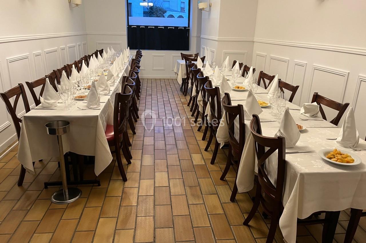 Salle de restaurant avec deux longues tables dressées, nappes blanches, serviettes pliées et assiettes de mise en bouche.