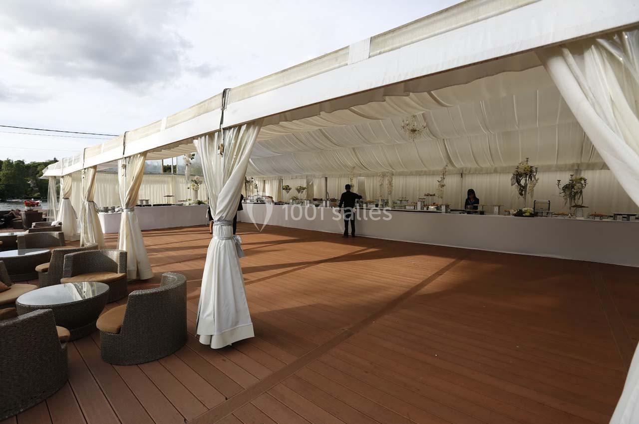 Tente blanche aménagée avec des tables de buffet et des fauteuils sur une terrasse en bois, en extérieur.