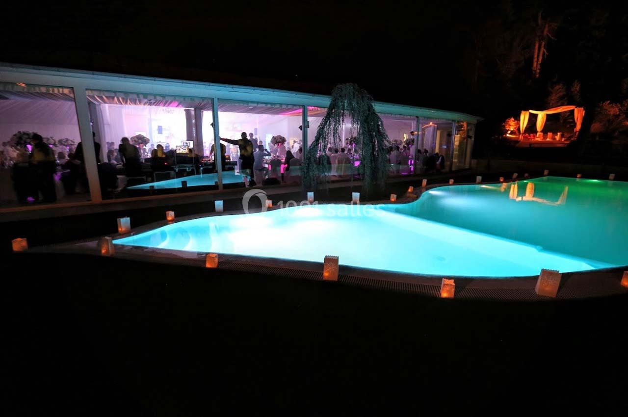Piscine éclairée de nuit entourée de bougies, avec une salle vitrée illuminée en arrière-plan.