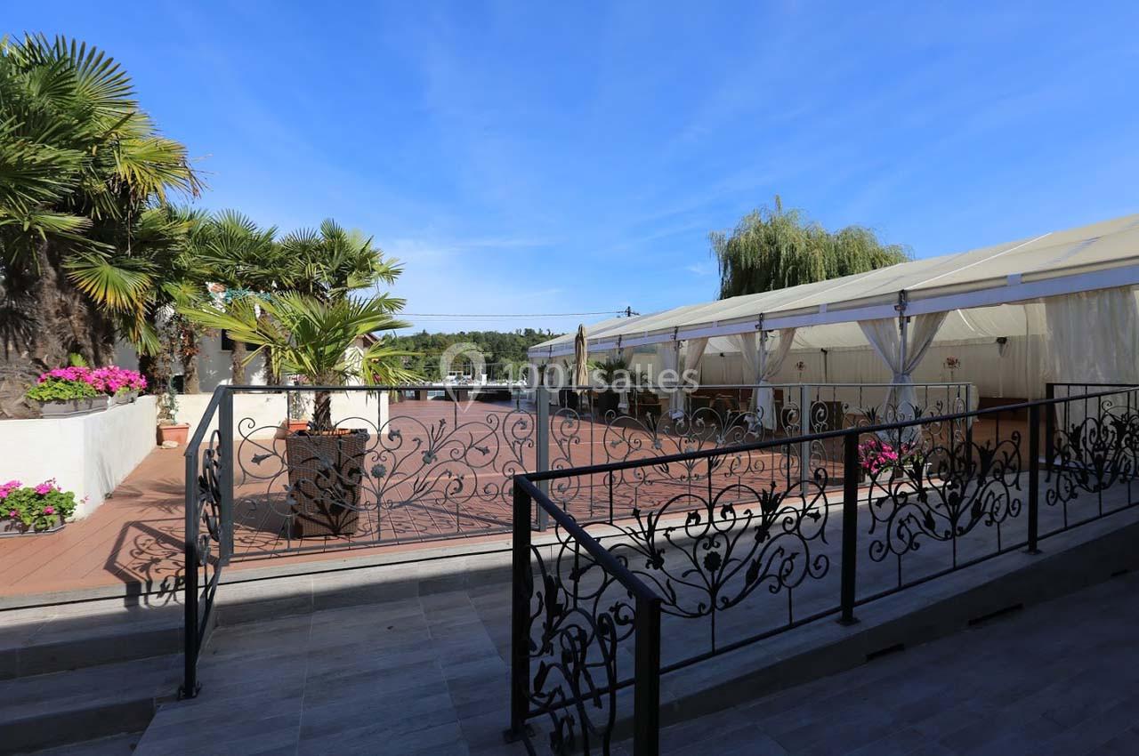 Terrasse extérieure avec sol en bois, rambarde en fer forgé, plantes en pot et tente blanche en arrière-plan.