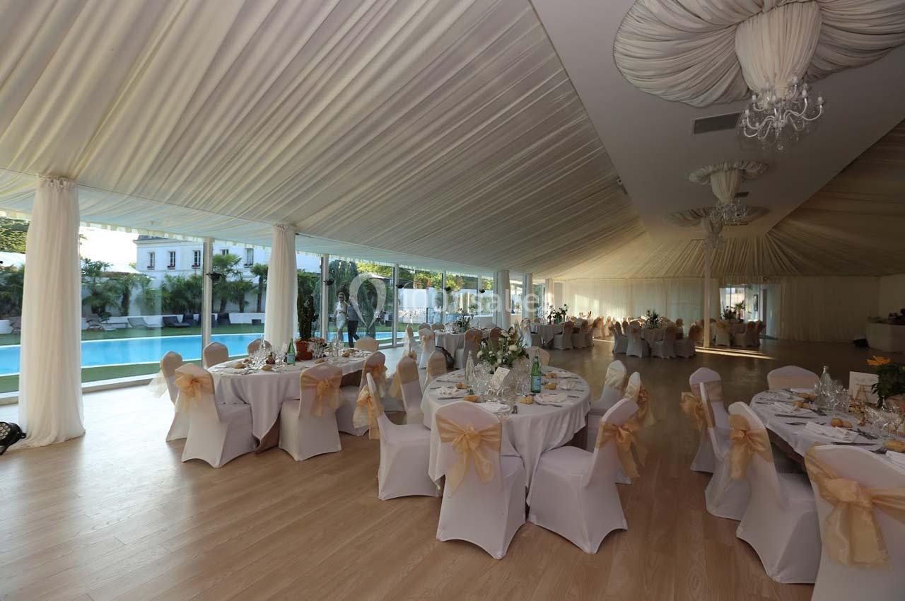 Salle de réception lumineuse avec tables rondes décorées, vue sur une piscine et un jardin à travers de grandes baies…
