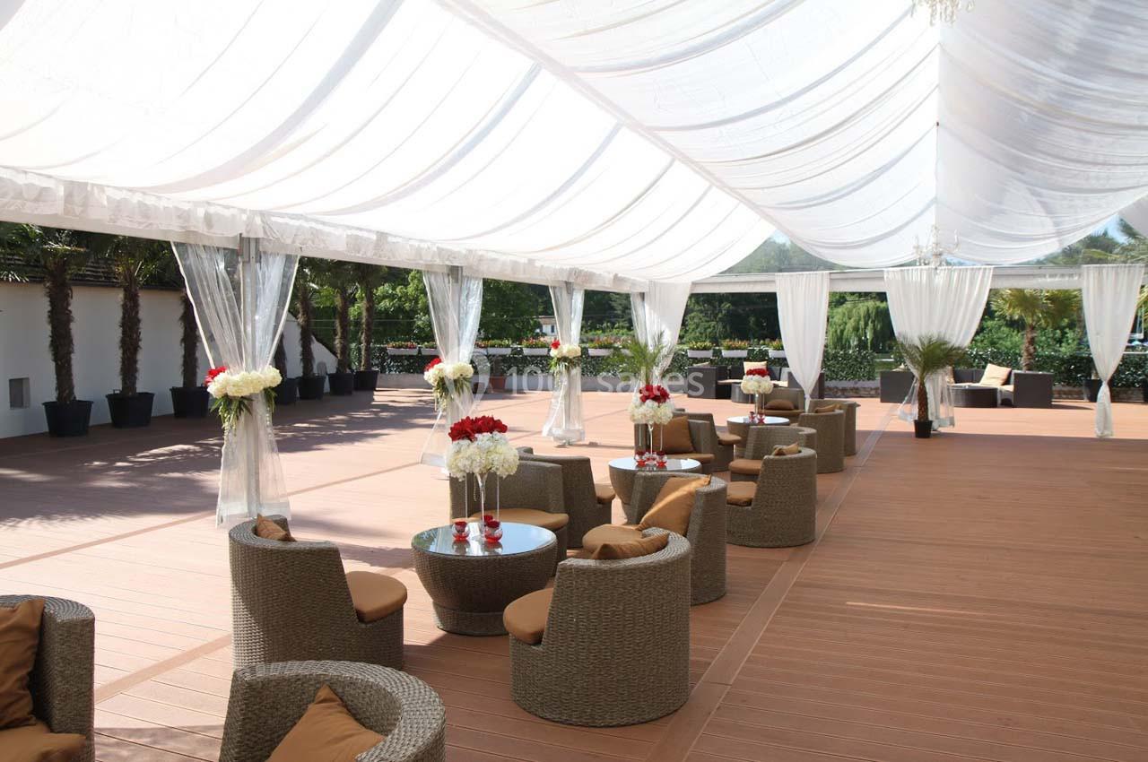 Espace extérieur couvert avec des fauteuils en rotin, tables basses et décoration florale sous une toile blanche translucide.