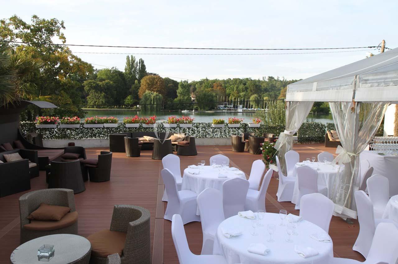 Terrasse aménagée avec tables dressées et fauteuils, offrant une vue sur un lac entouré de verdure et de fleurs.