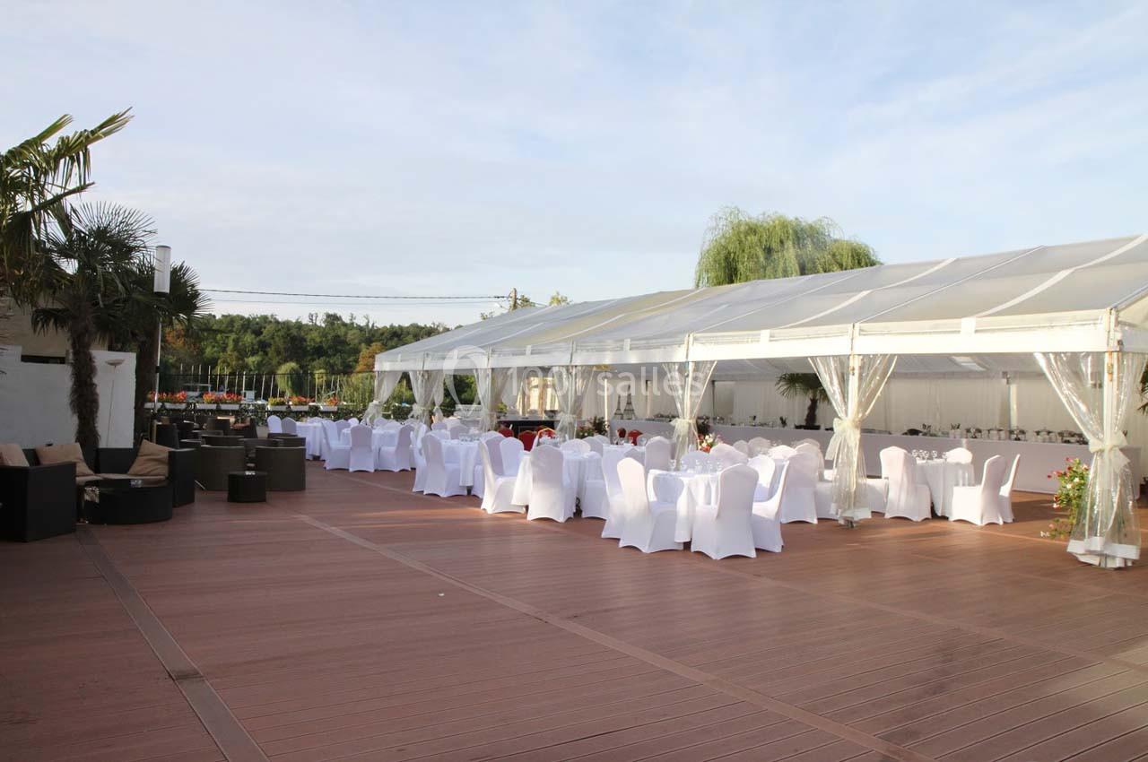 Grande terrasse en bois avec une tente blanche transparente abritant des tables rondes dressées et des chaises blanches.