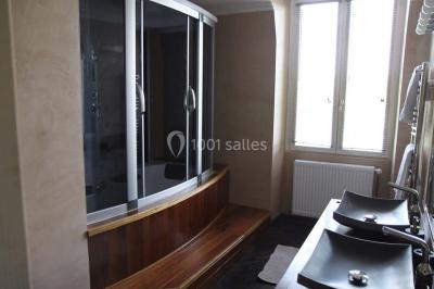 Salle de bain avec une cabine de douche moderne, un banc en bois intégré, deux vasques noires et une fenêtre avec stores.