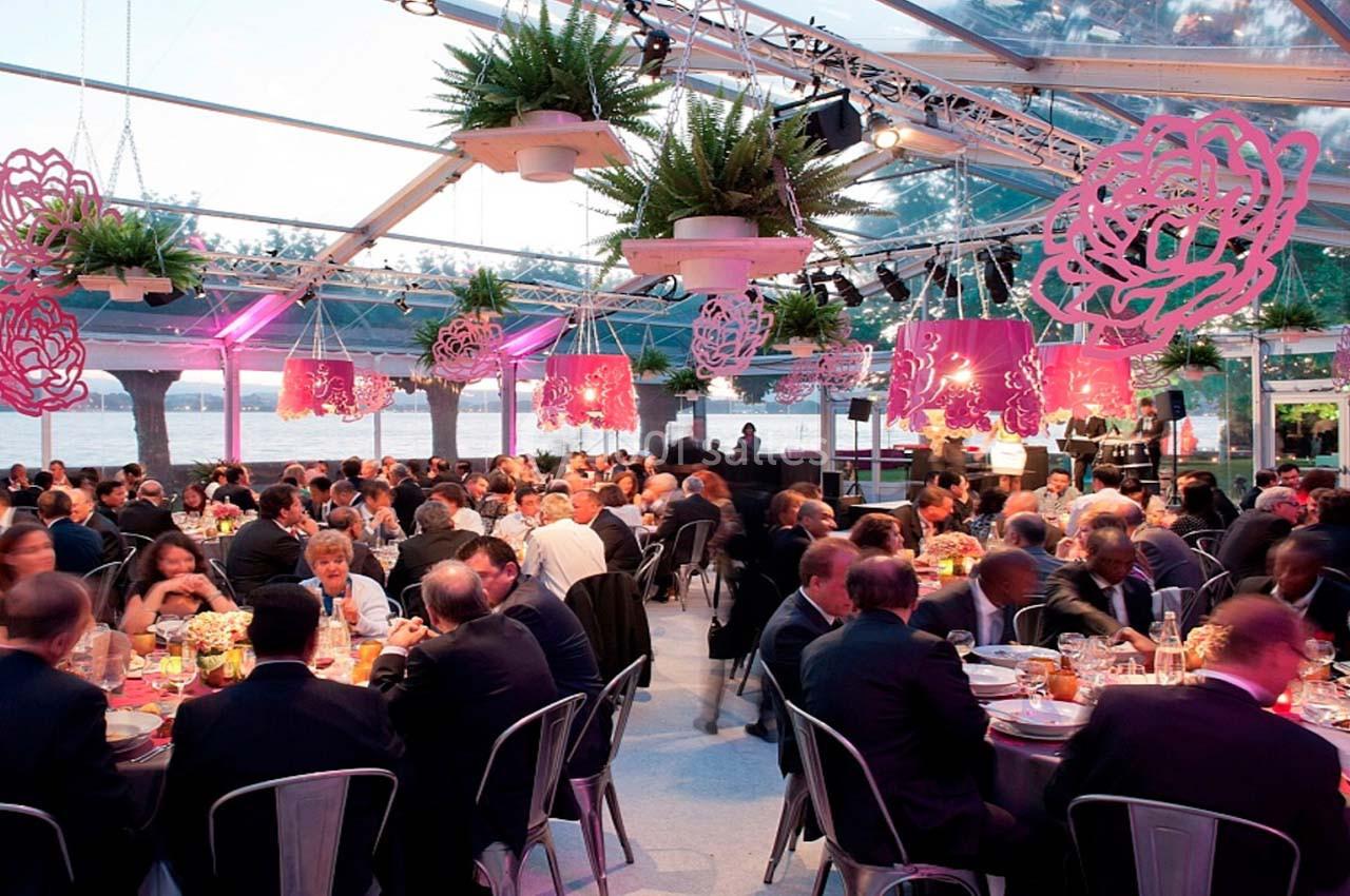 Salle de réception sous une verrière, avec des invités assis à des tables décorées et une scène en arrière-plan.