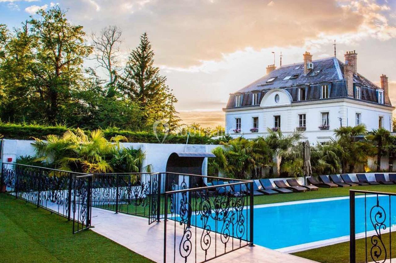 Piscine extérieure entourée de transats et de végétation, avec une grande maison en arrière-plan sous un ciel ensoleillé.