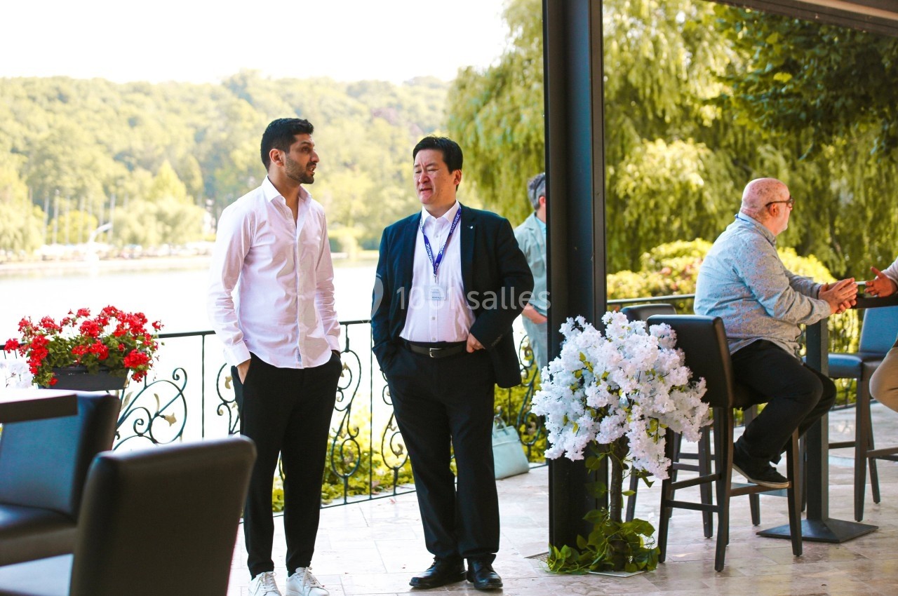 Deux hommes discutent debout sur une terrasse avec vue sur un lac et des fleurs blanches en décoration.