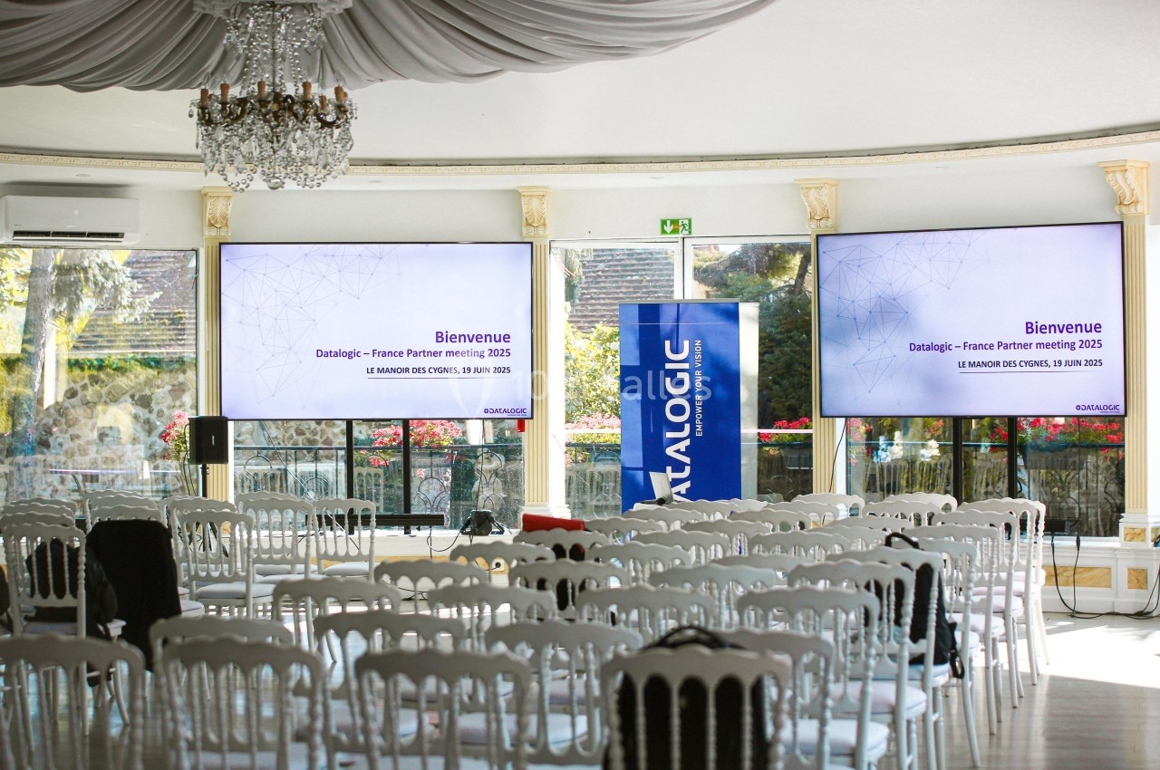 Salle de conférence lumineuse avec des chaises blanches alignées, deux écrans affichant ’Bienvenue’ et une bannière.