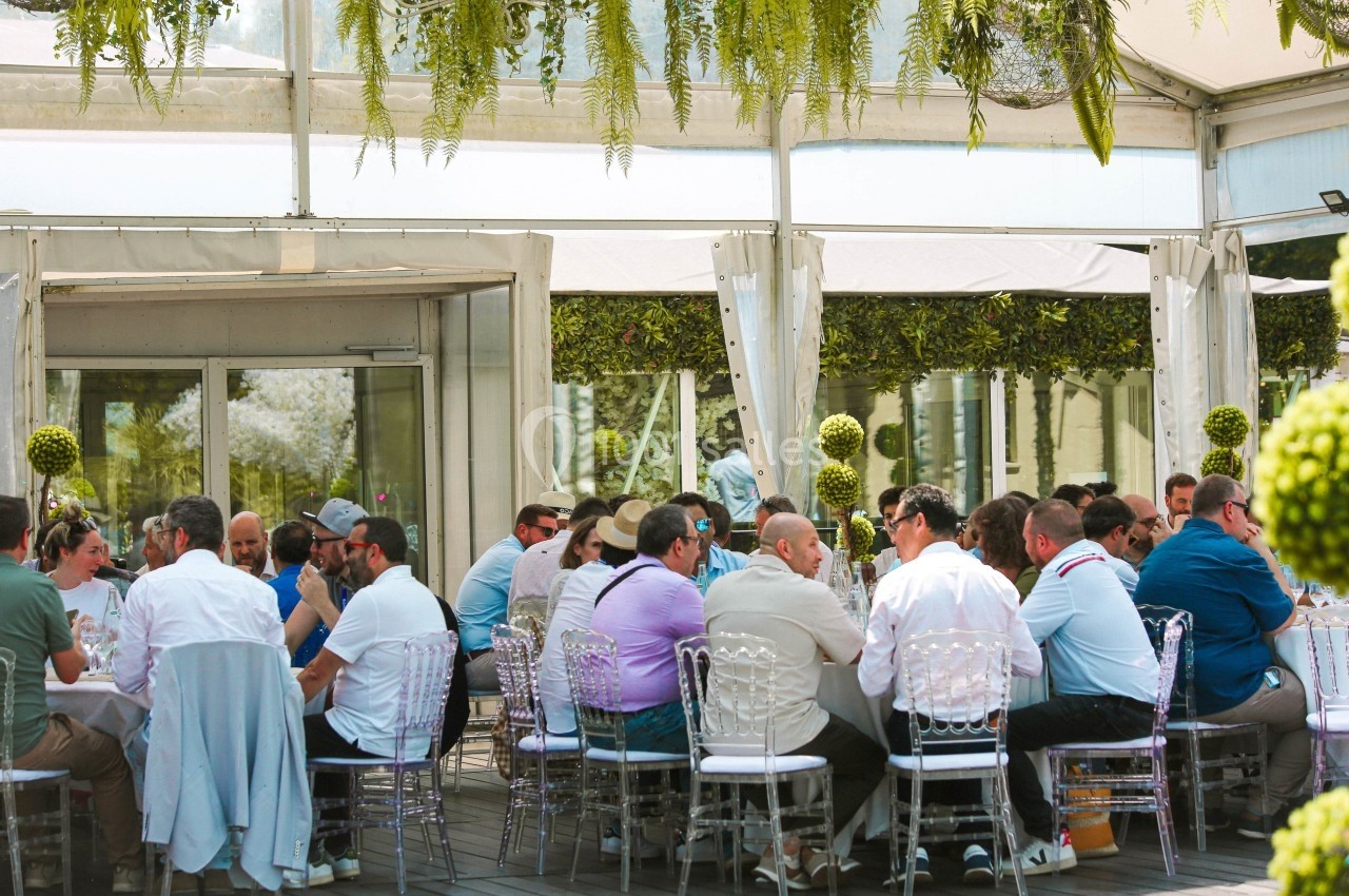 Groupe de personnes assises autour de tables lors d'un événement en extérieur sous une structure couverte.