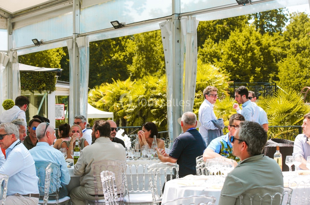 Des personnes assises et debout discutent lors d'un événement en plein air sous une tente, entourées de végétation.