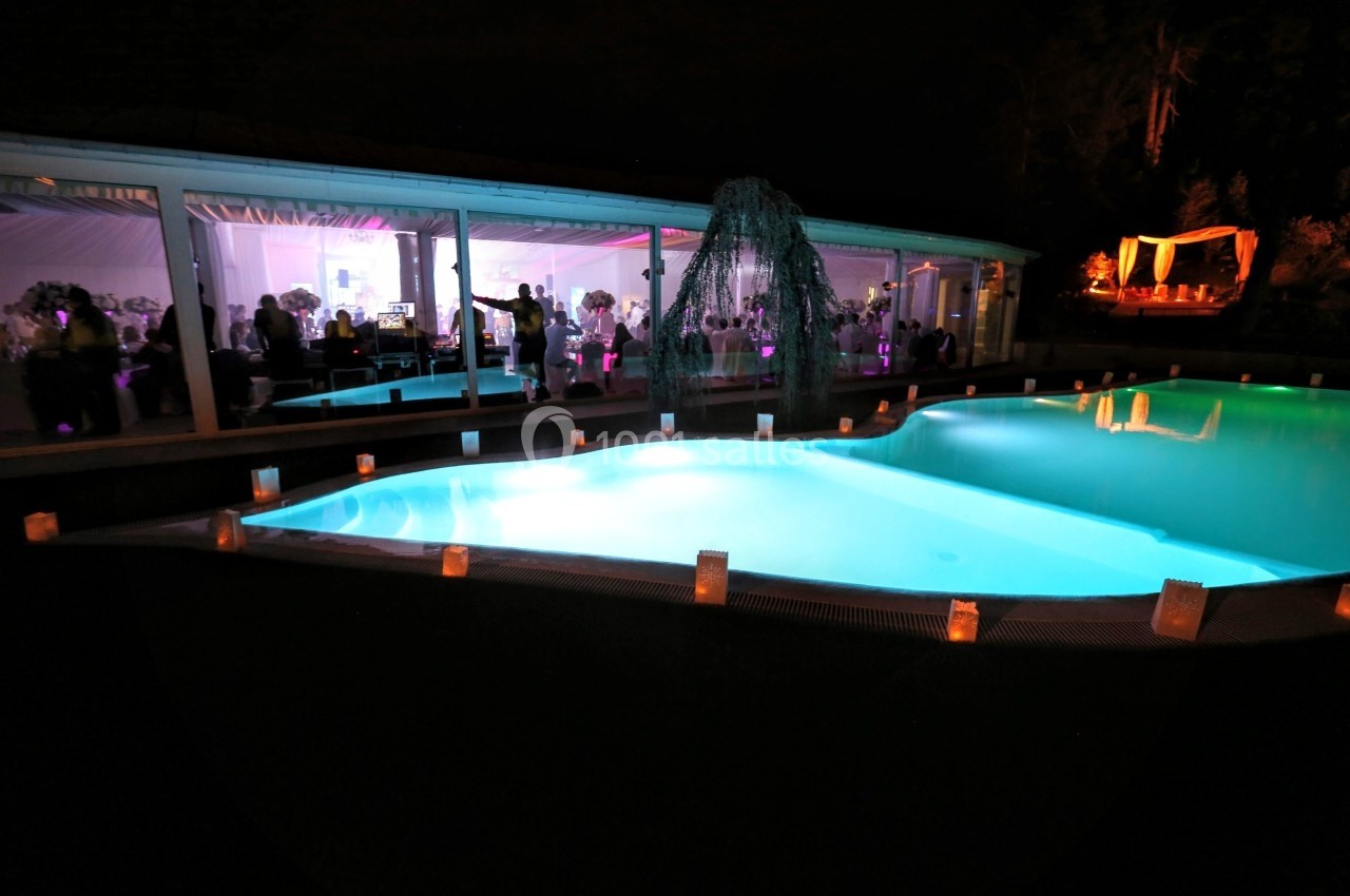 Piscine éclairée de nuit entourée de bougies, avec une salle vitrée illuminée en arrière-plan.