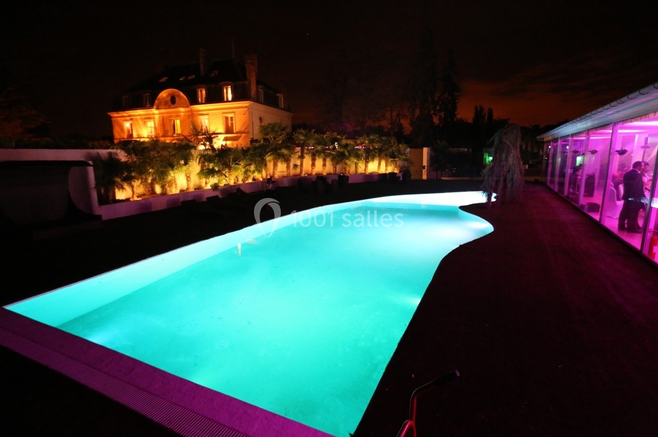 Piscine éclairée de nuit devant une grande maison illuminée et une terrasse avec lumières colorées.