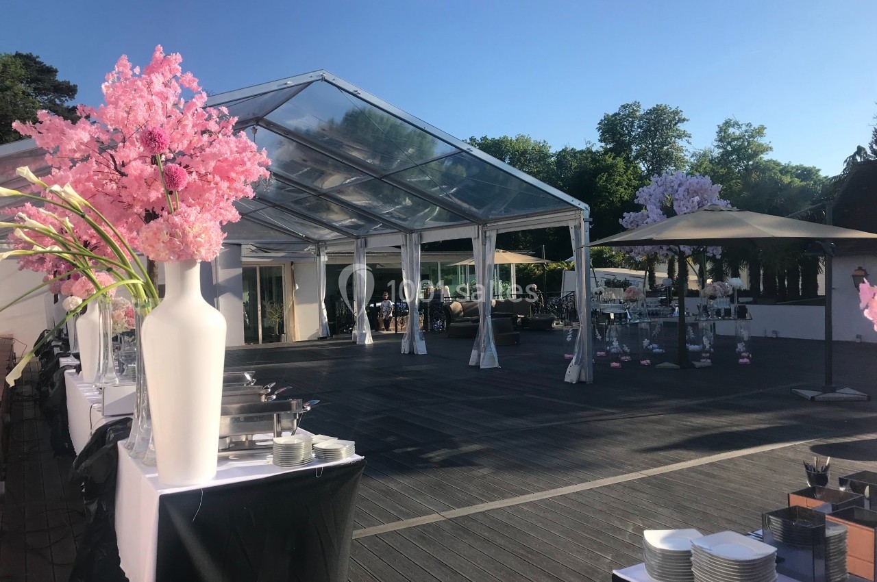 Structure en plein air avec toit transparent, tables dressées, décorations florales et ciel dégagé en arrière-plan.