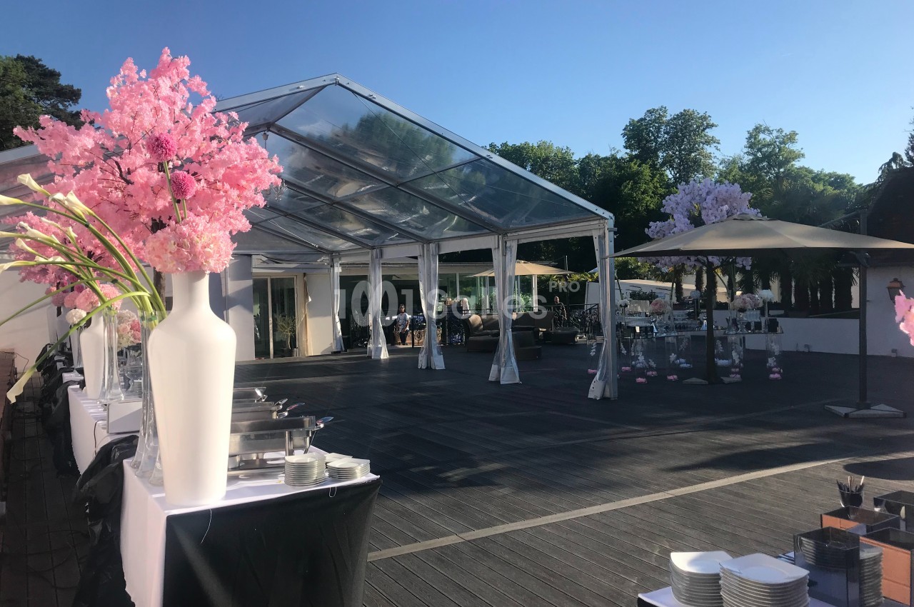 Structure en plein air avec toit transparent, tables dressées, décorations florales et ciel dégagé en arrière-plan.
