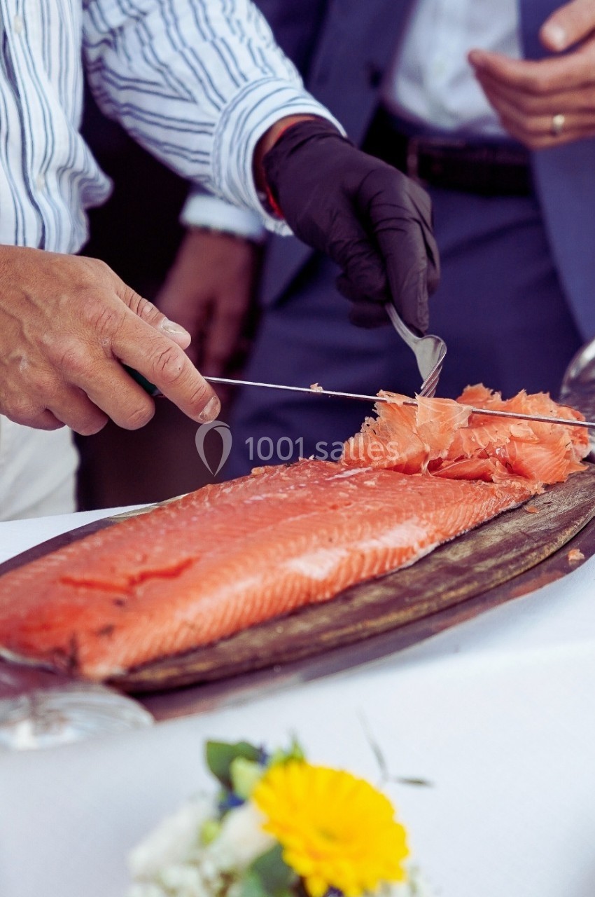 Un homme tranche un filet de saumon frais posé sur une planche en bois, avec une table décorée en arrière-plan.