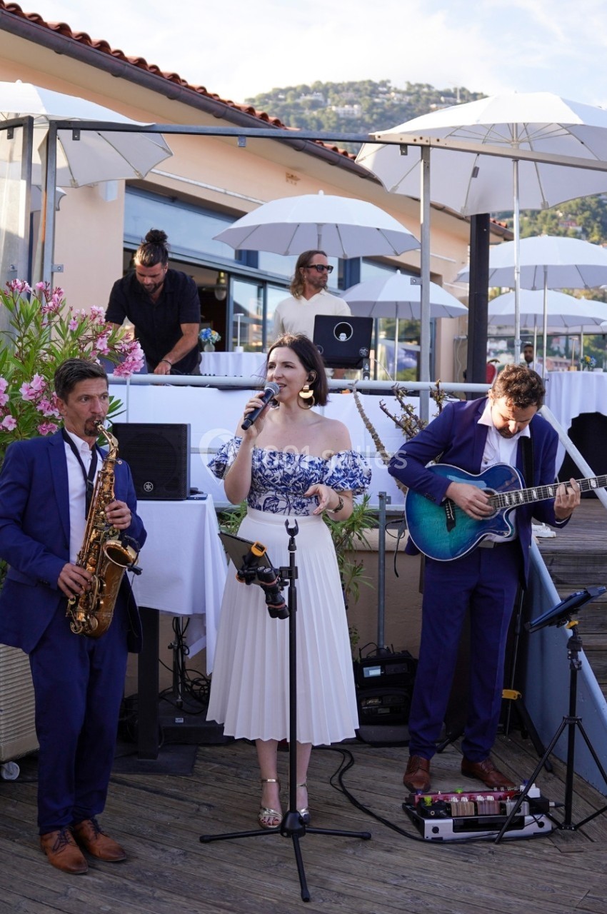 Un groupe de musiciens joue en plein air sur une terrasse, avec une chanteuse, un saxophoniste et un guitariste.