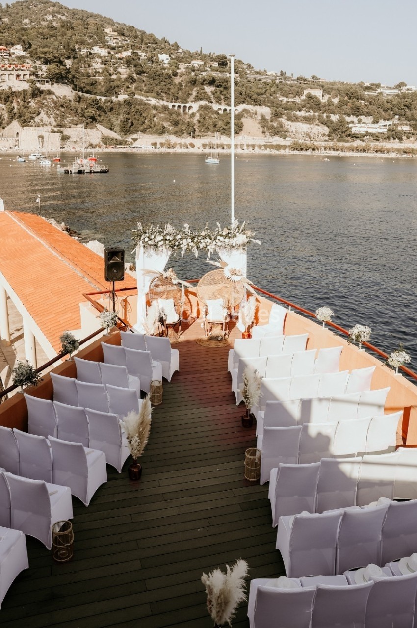 Chaises blanches alignées sur le pont d'un bateau, décoré pour une cérémonie, avec vue sur la mer et les collines.