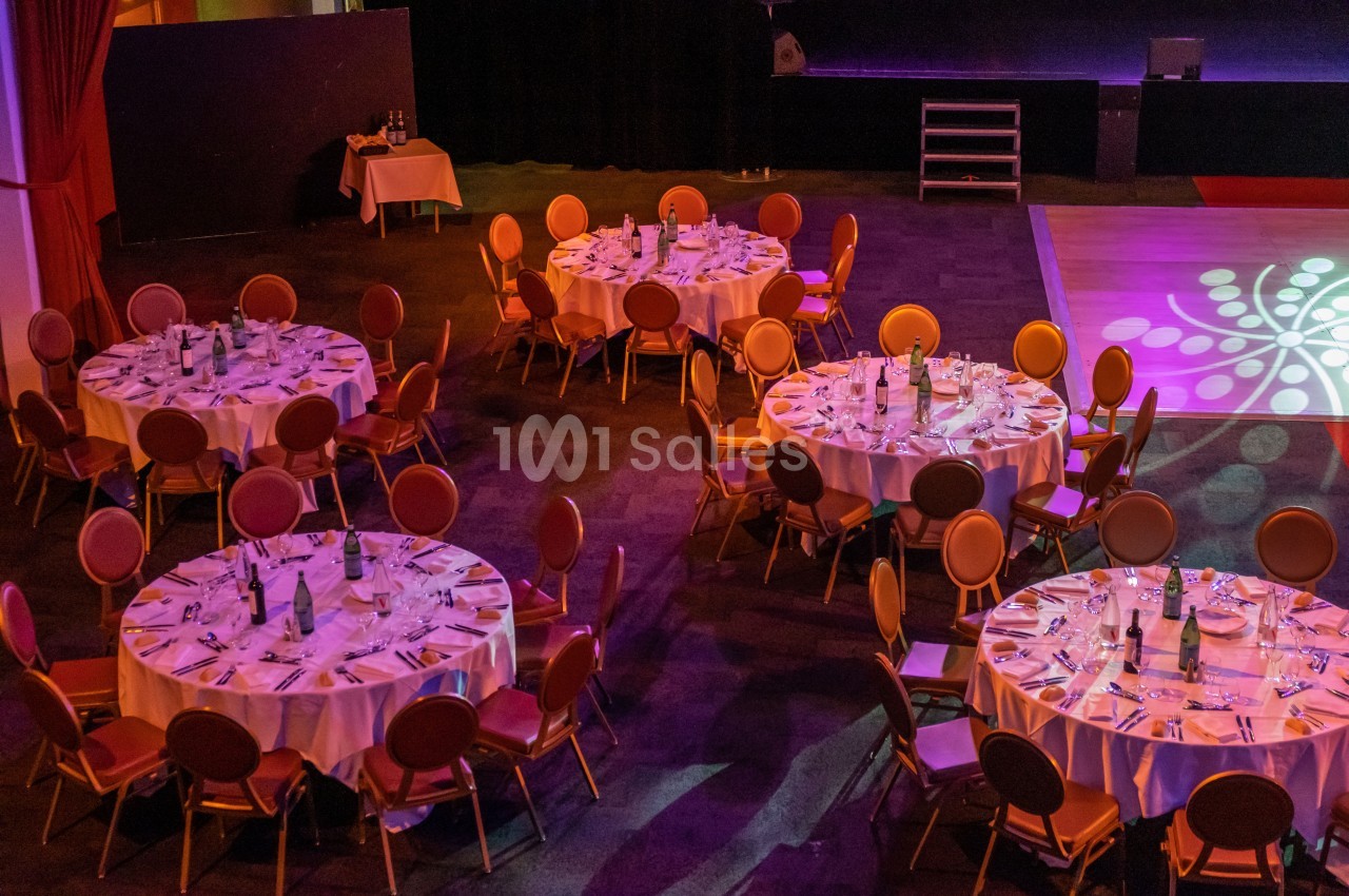 Salle de réception avec tables rondes dressées, nappes blanches, chaises disposées et éclairage coloré.
