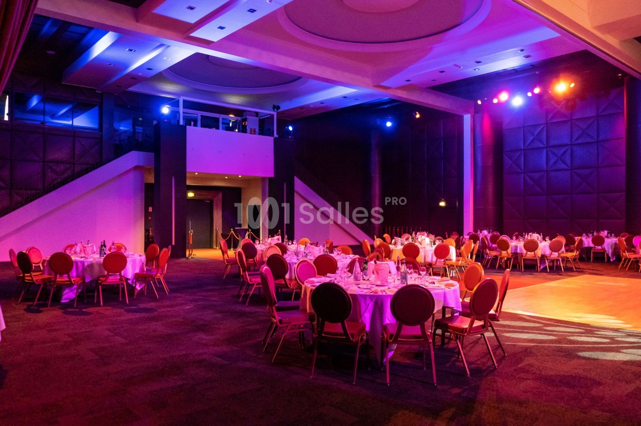 Salle de réception avec tables rondes dressées, éclairage coloré et scène illuminée en arrière-plan.