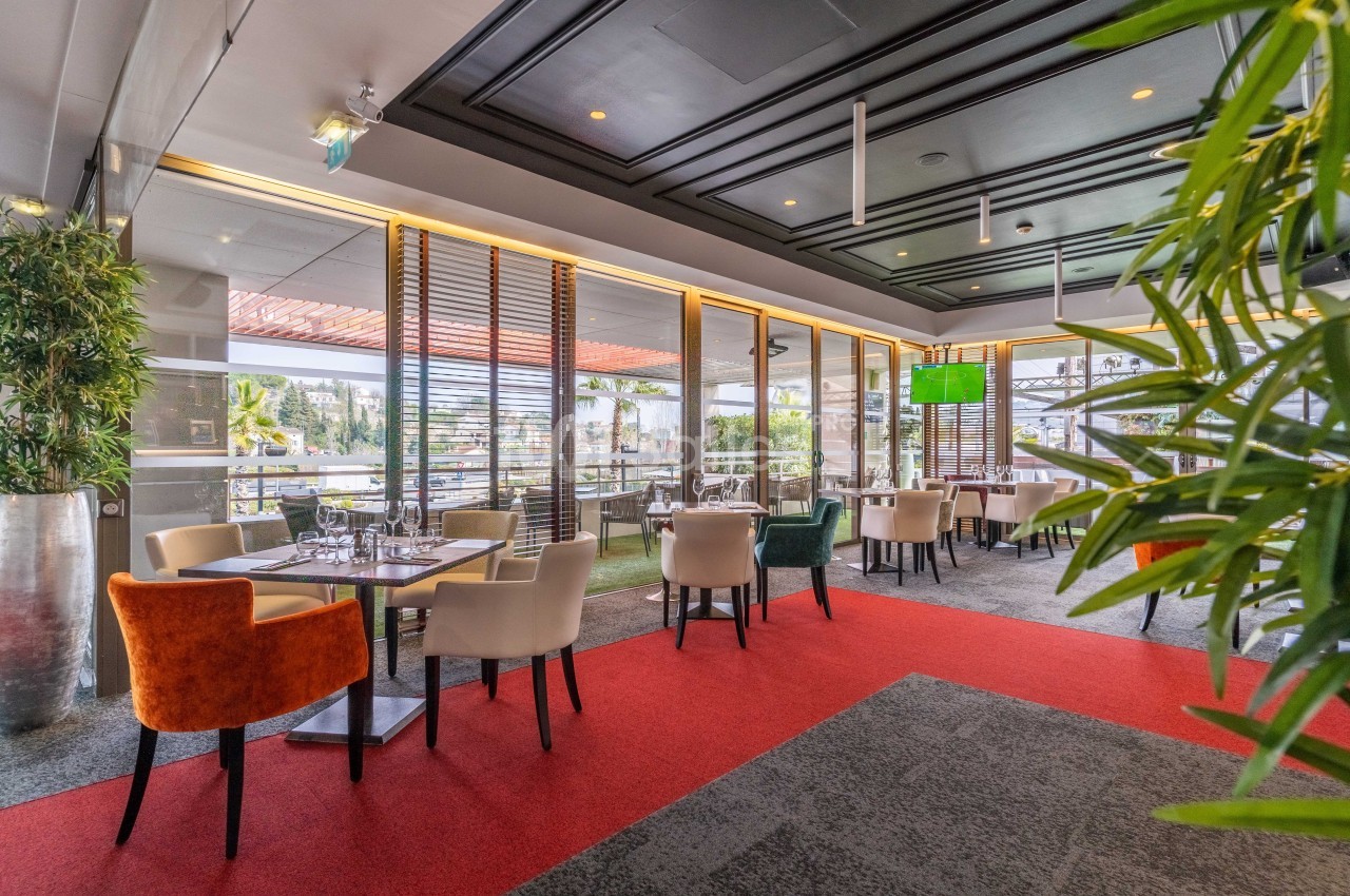 Salle de restaurant lumineuse avec grandes baies vitrées, tables dressées et chaises colorées, vue sur l'extérieur.