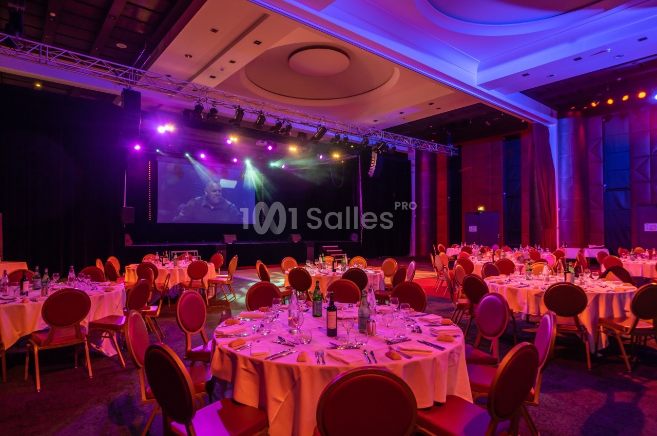 Salle de réception avec tables dressées, éclairage coloré et scène équipée d'un écran géant en arrière-plan.