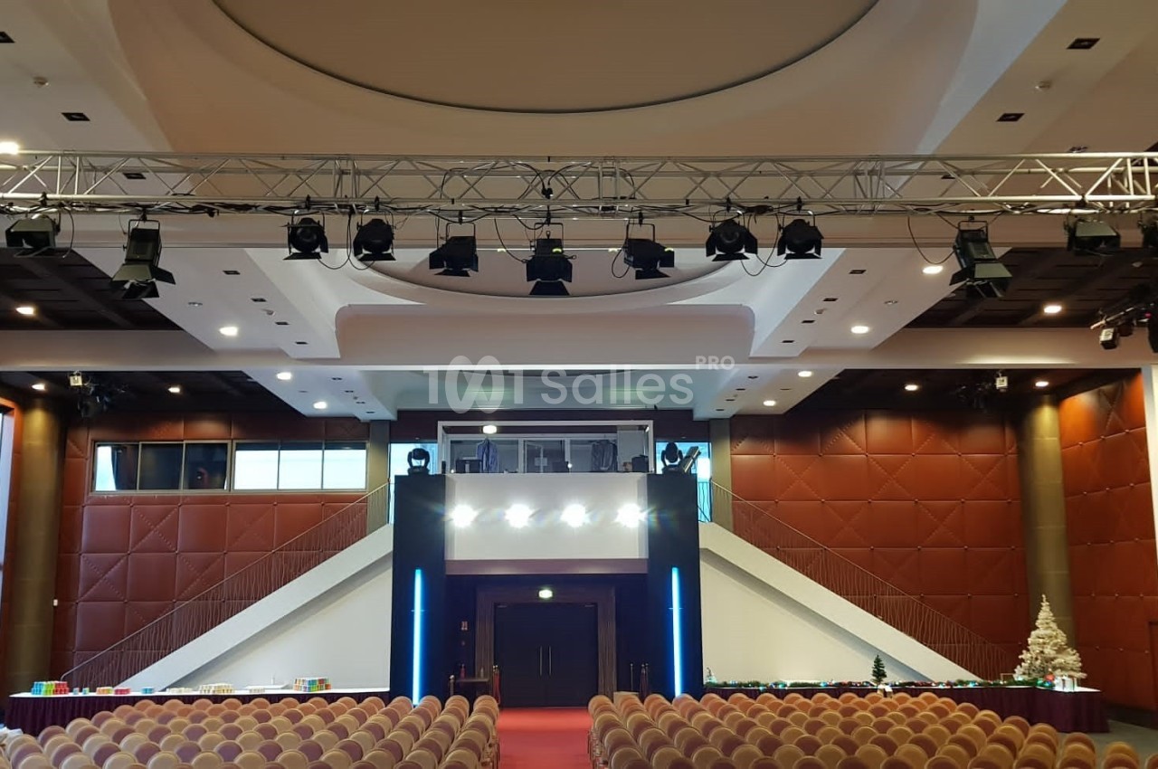 Salle de conférence avec sièges alignés, scène centrale, éclairages suspendus et sapin décoratif à droite.