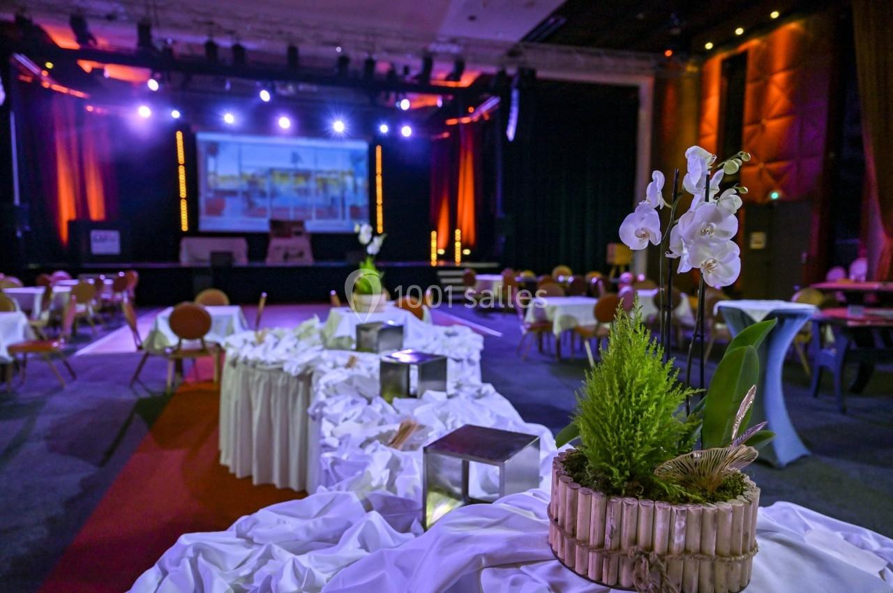 Salle de réception décorée avec des tables drapées de blanc, des plantes et un éclairage coloré en arrière-plan.