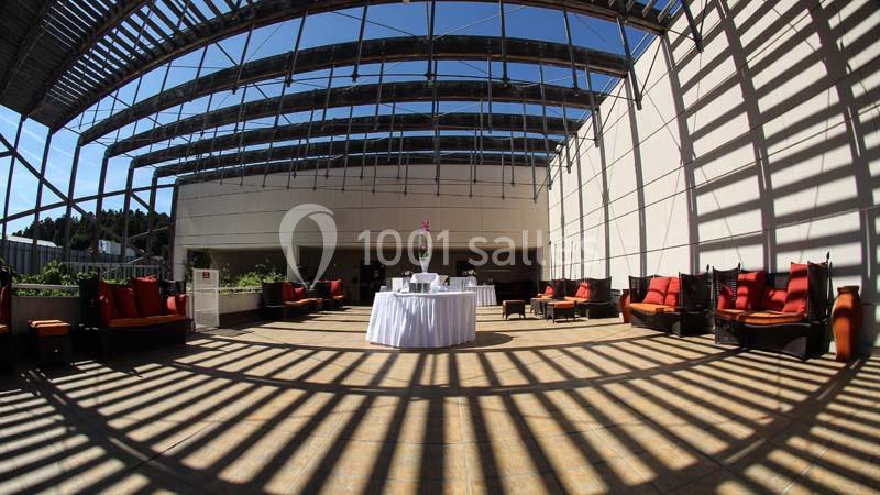 Salle lumineuse avec toit en verre, tables dressées au centre et fauteuils rouges disposés le long des murs.
