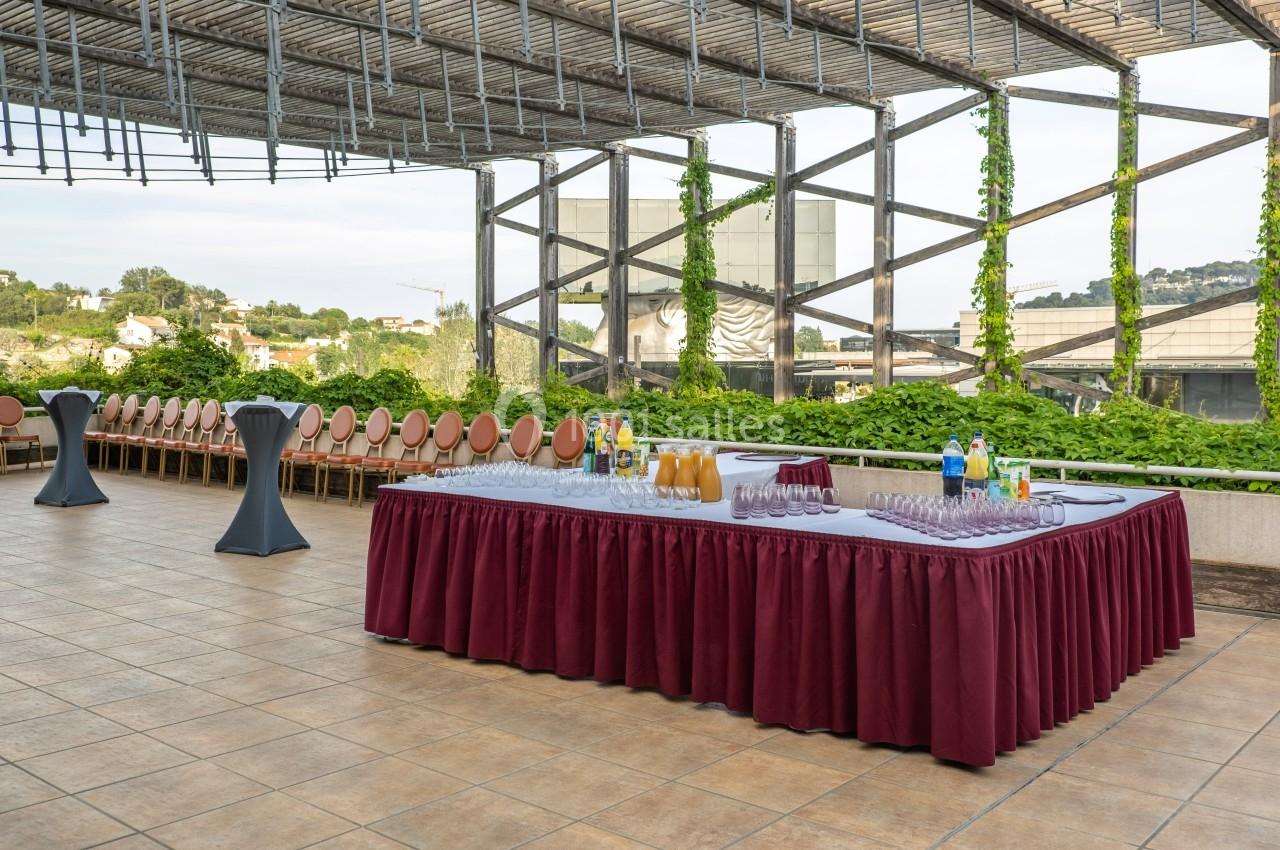 Table de réception dressée avec boissons et verres sur une terrasse extérieure entourée de verdure.