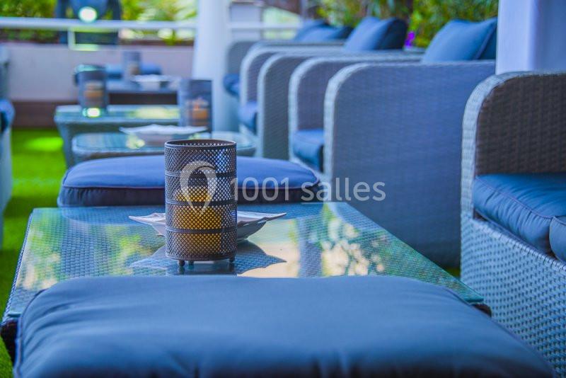 Mobilier de jardin avec fauteuils en rotin gris, coussins bleus et tables en verre, décoré de bougies dans des photophores.