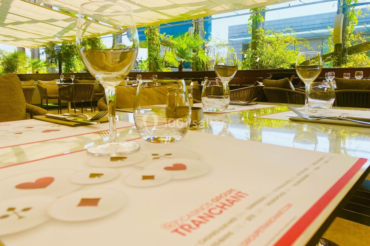 Table dressée en terrasse ensoleillée avec des verres, des couverts et des cartes à jouer imprimées sur le menu.